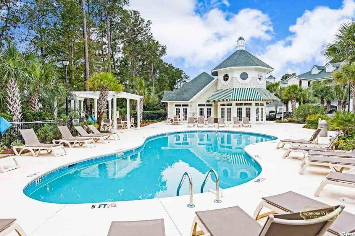 A sparkling outdoor swimming pool surrounded by lounge chairs and shaded by swaying palm trees. In the background, the clubhouse is visible, creating an inviting space for relaxation and social gatherings under the sun.