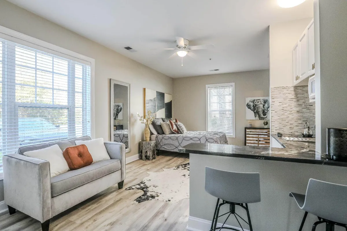 A tastefully decorated studio with a bed adorned with decorative pillows, a sofa with an accent pillow, an artful wall, and a kitchenette area visible in the background. The layout encourages easy socializing, relaxation, and meal preparation in a stylish setting.