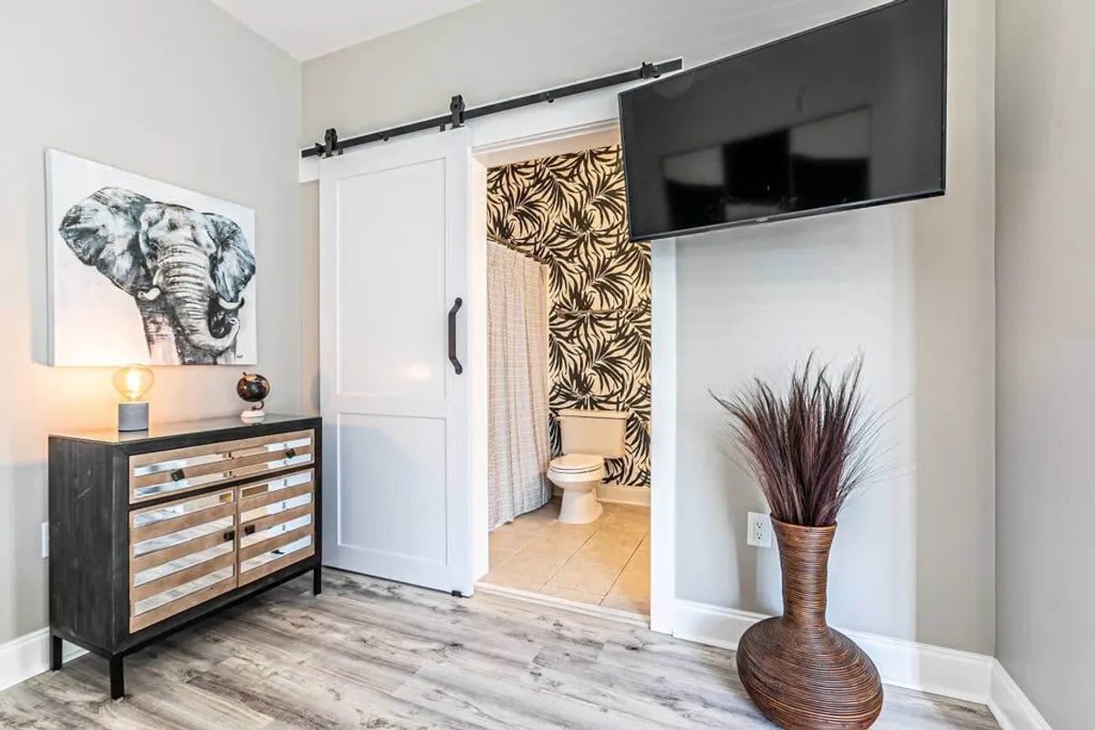 A stylish, modern suite interior featuring a sliding barn door that opens toward a bathroom, with a sleek modern dresser, flat-screen TV, and decorative vase. The angle suggests the bathroom is a private, accessible retreat off the main living space, emphasizing a sense of privacy and relaxation.