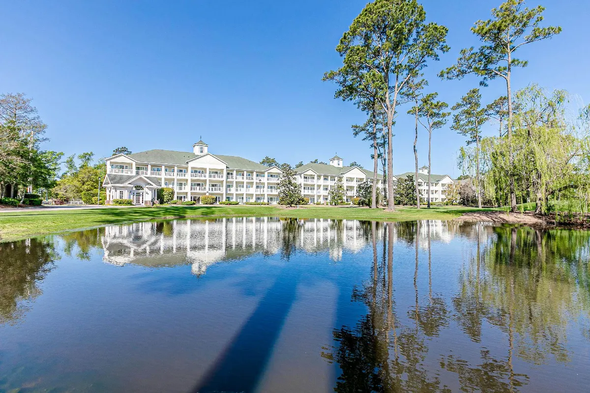 A serene scene featuring tranquil waters reflecting the building’s architecture, framed by tall trees. The open lawn and peaceful surroundings evoke a sense of relaxation and the opportunity for outdoor leisure activities such as strolls, picnics, or simply unwinding by the lake.