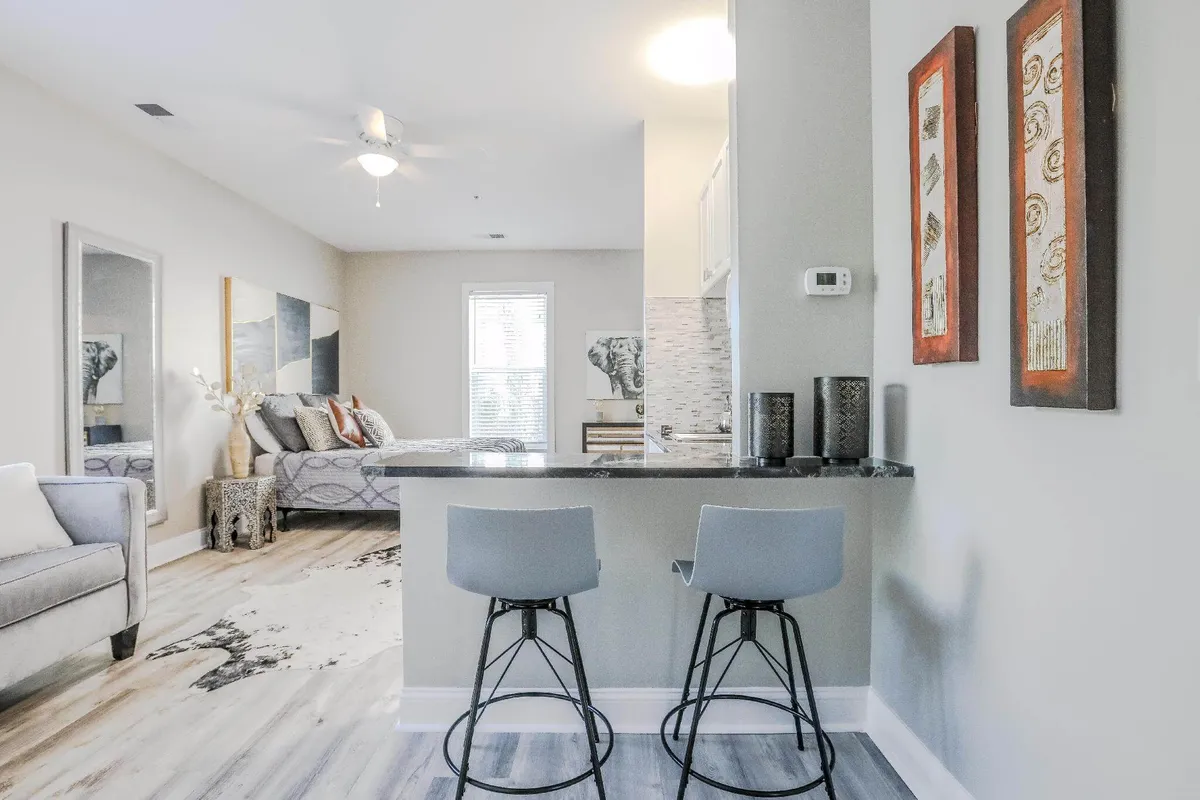The image displays a contemporary studio layout with a kitchen island, seating area, and a visible bedroom area. Decorative wall art and modern furnishings give the space a cozy, welcoming feel. Natural light streams in through windows, emphasizing the studio's airy and warm ambiance.
