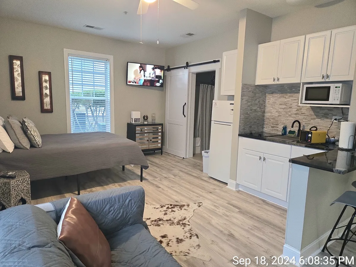 A stylish, open-plan studio with a plush bed, comfortable sofa, wall-mounted TV, and a compact yet well-equipped kitchenette. Natural light streams through a nearby window, creating a bright and inviting environment perfect for relaxation or a cozy meal.