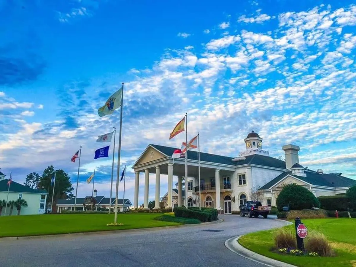 A grand entrance with stately columns, multiple flags flying, and a meticulously maintained lawn framed by decorative landscaping. The impressive exterior sets the tone for arrival and welcomes guests, hinting at the upscale amenities within.