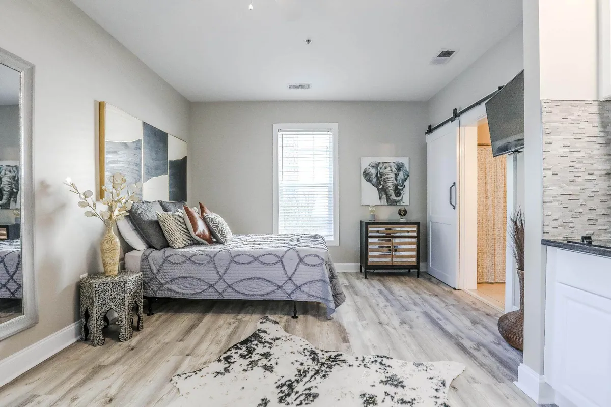 The image showcases a serene and stylish interior studio space featuring a cozy bed adorned with decorative boho pillows, a large mirror that opens up the room, and striking elephant artwork above a chic nightstand. Soft natural light filters in, creating a peaceful and inviting ambiance.