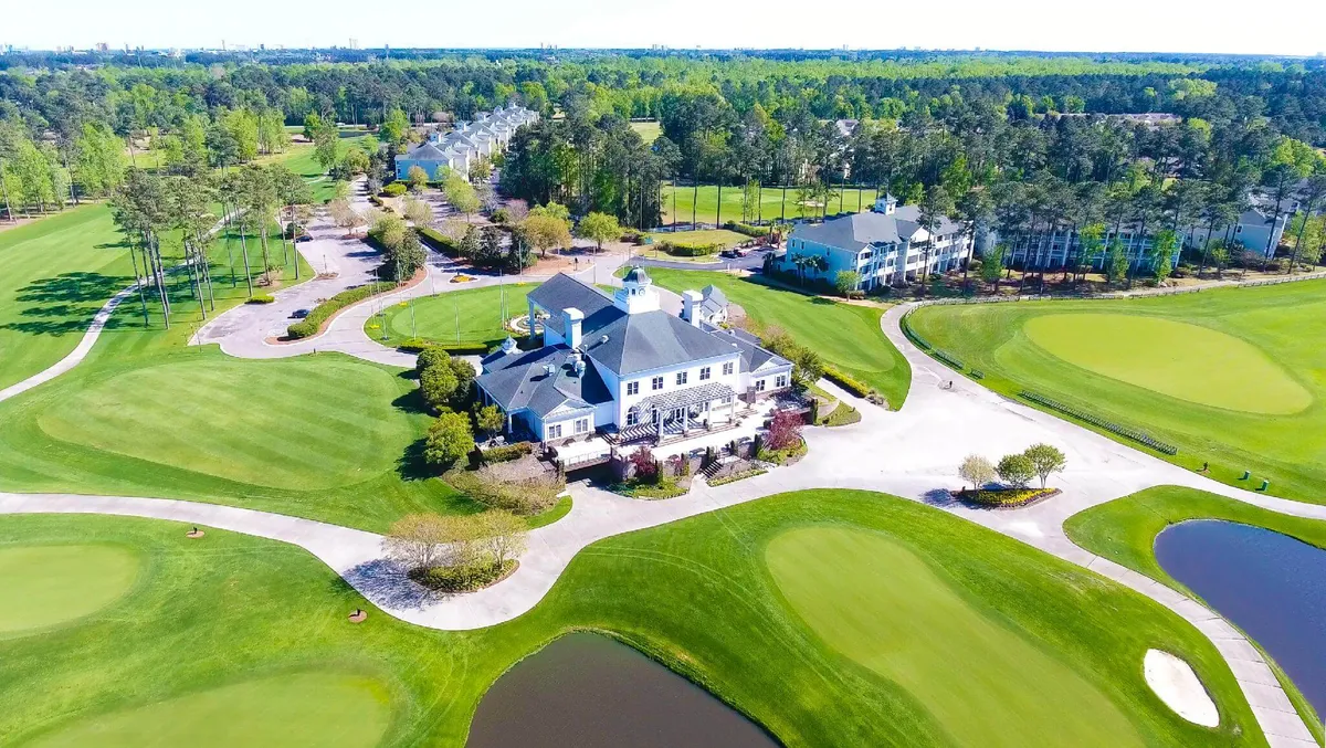 A picturesque exterior view of the golf clubhouse flanked by vast lawns, a glistening pond, and mature trees. The clubhouse’s inviting patio hints at post-game social gatherings or relaxed outdoor dining, with the course stretching beyond.