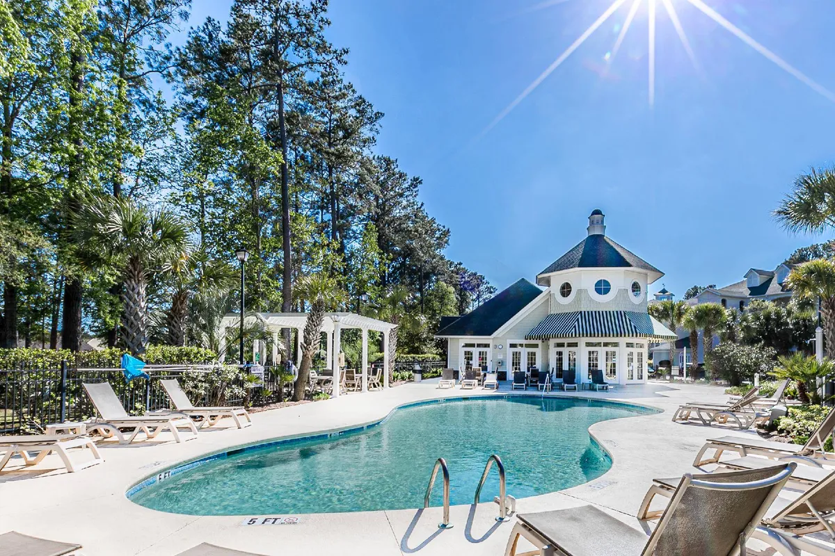 This image shows the resort’s sparkling swimming pool surrounded by lounge chairs and shaded by mature trees. The elegant clubhouse is visible in the background, tying the pool area to the resort’s social hub. The inviting water and sunny setting suggest an ideal spot for relaxation or social gatherings.