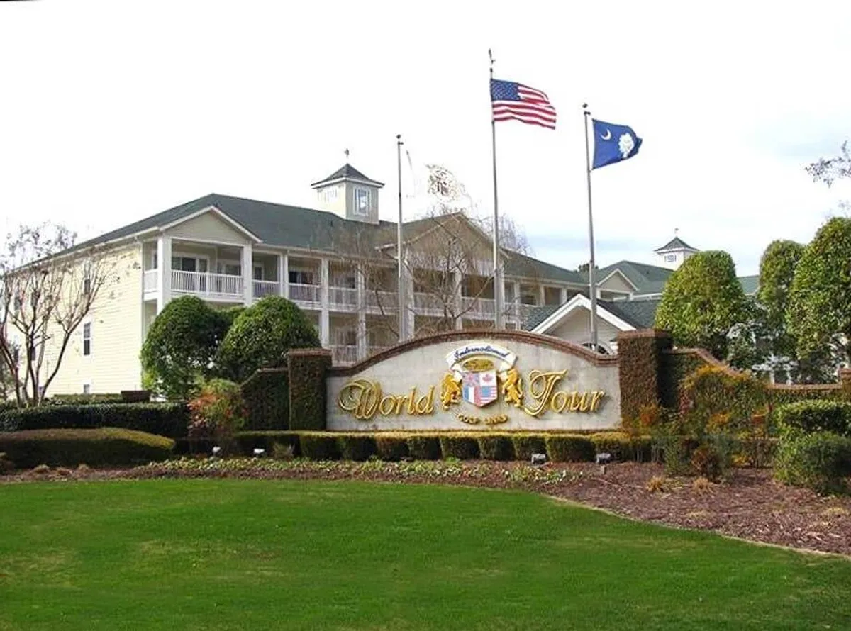 The welcoming World Tour sign, surrounded by flags and beautifully landscaped gardens, stands at the entrance of the resort. The building facade in the background provides a sense of grandeur and arrival.